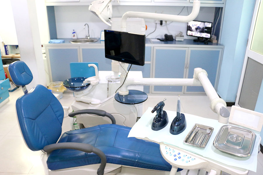 Bright dental treatment room with a blue dental chair, monitor, treatment light, instruments, and pale blue cabinetry